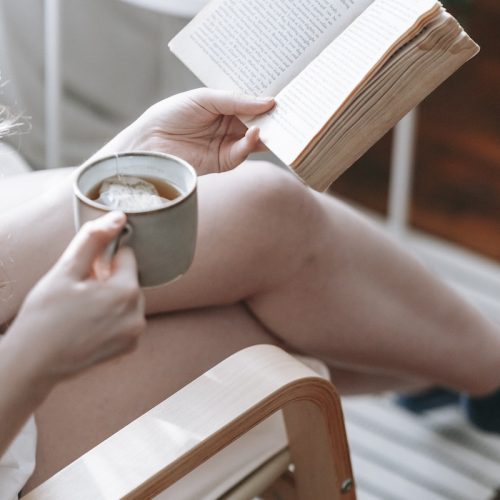 From above of crop anonymous female sitting with crossed legs and drinking tea while reading book at weekend